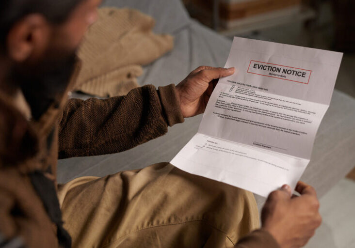 Person holding eviction notice while sitting on couch and reading in a dimly lit room without natural light coming in through the window behind