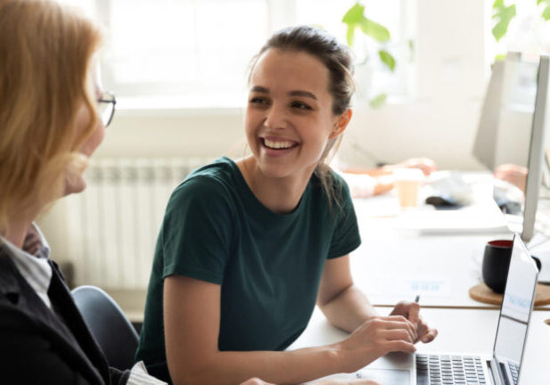 Pleasant workday. Two women colleagues employees of diverse age sharing points of view at workplace in office, happy smiling millennial female trainee discussing business ideas with adult aged trainer