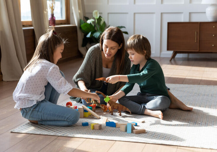 Caring mom or nanny sit on floor play with building bricks toys enjoy weekend with little kids, loving happy young mother have fun engaged in funny activity with small children in living room